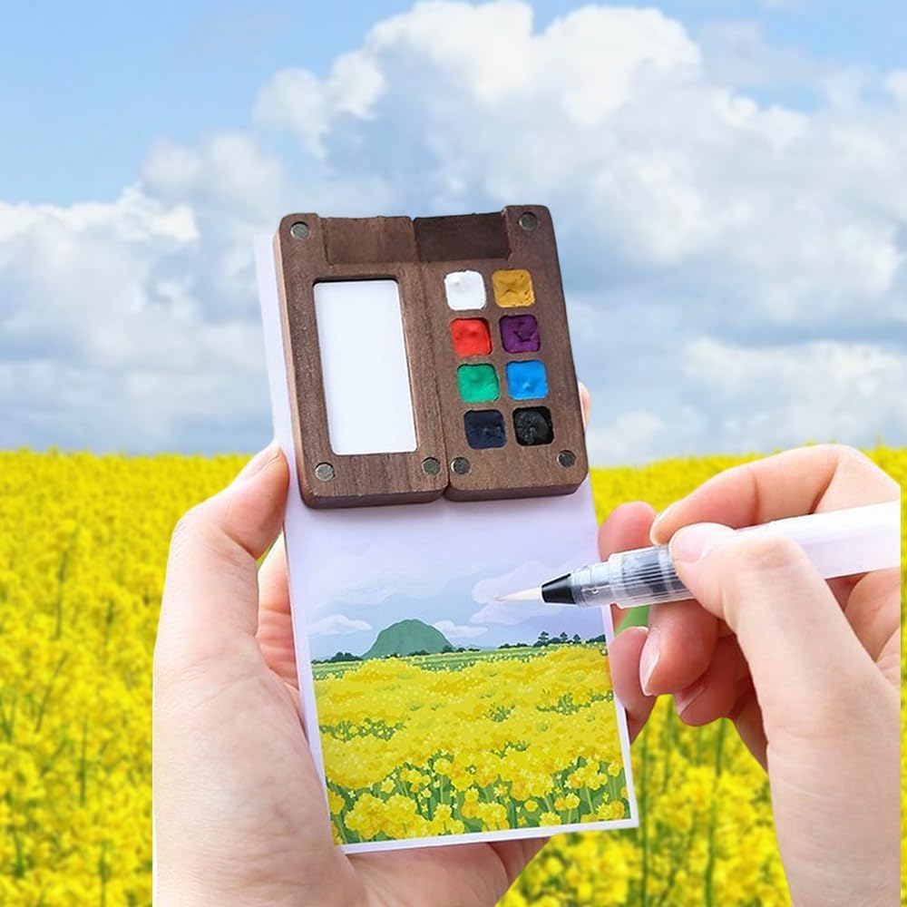 Hand holding a small watercolor palette with a field of yellow flowers and blue sky in the background